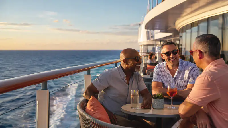Three LGBTQ+ friends talking on a modern cruise ship deck at golden-hour sail-away