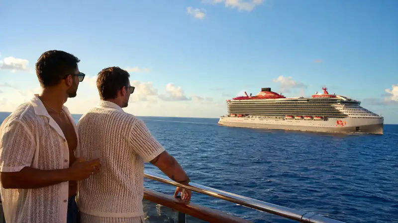 Two gay travelers watch a Virgin Voyages cruise ship from a sunny deck