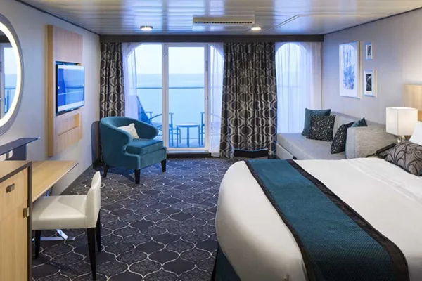 A cruise-ship balcony stateroom includes a bed, sofa, desk, and ocean view.