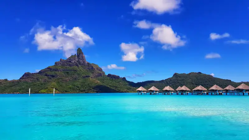 Overwater bungalows sit in a turquoise lagoon beneath a jagged tropical island.