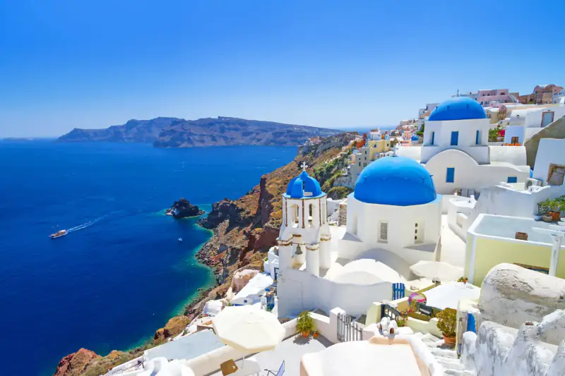 White buildings cling to a rocky Greek island above a blue sea.