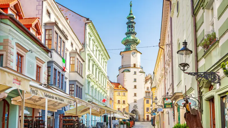 A narrow European street leads toward a clock tower and church spire.