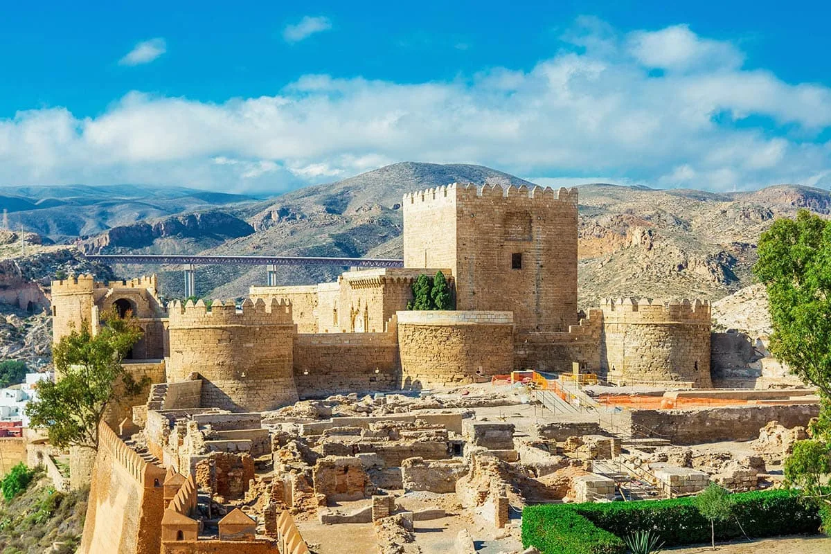 A stone kasbah stands in a warm desert landscape beneath Atlas Mountains.