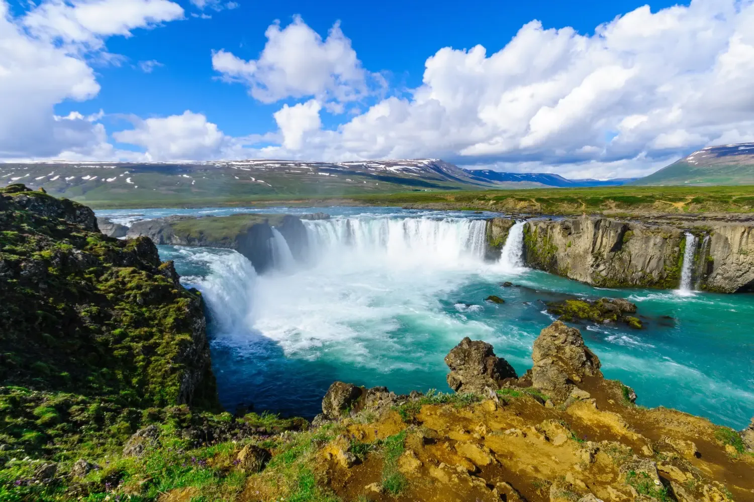 A waterfall drops through a green Icelandic valley beneath a snow-capped mountain.