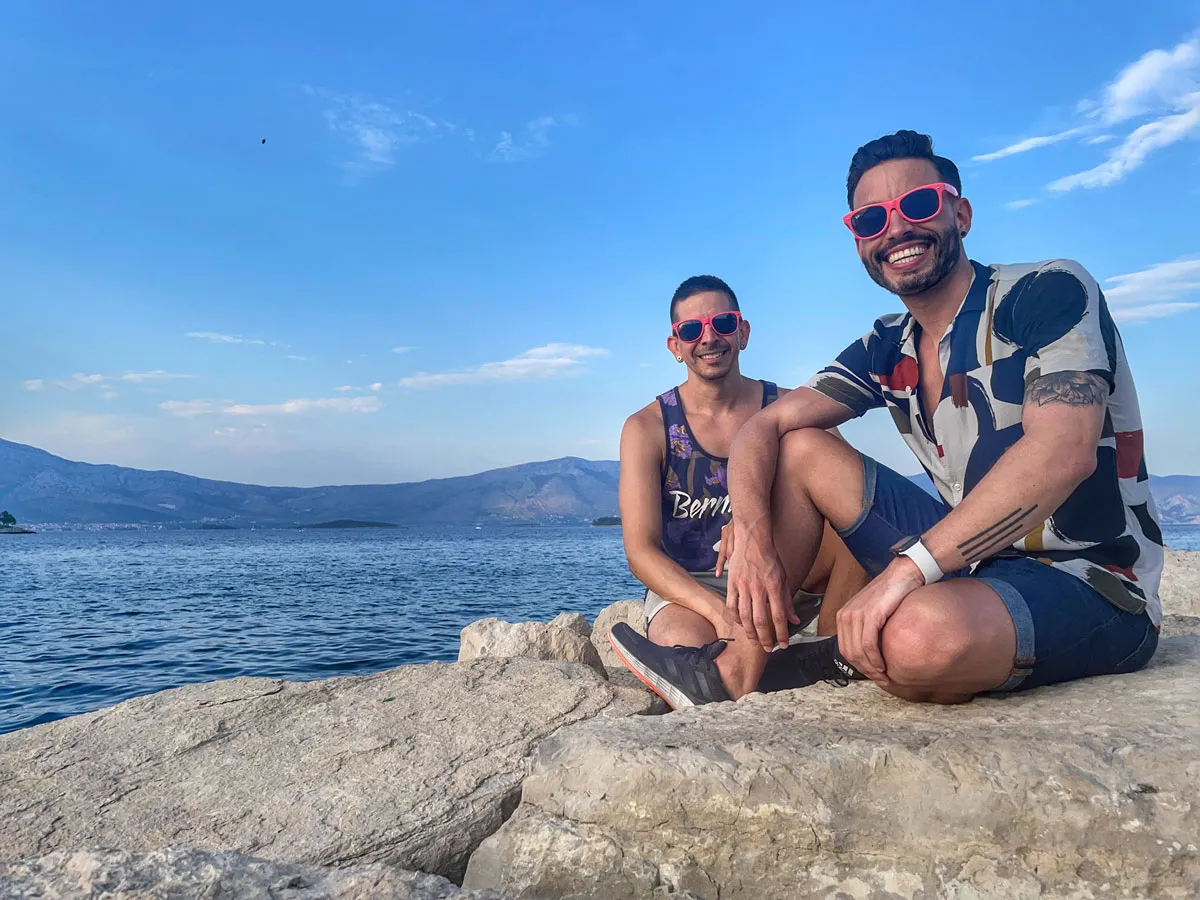 Two smiling travelers sit beside bright blue water on the Croatian coast.